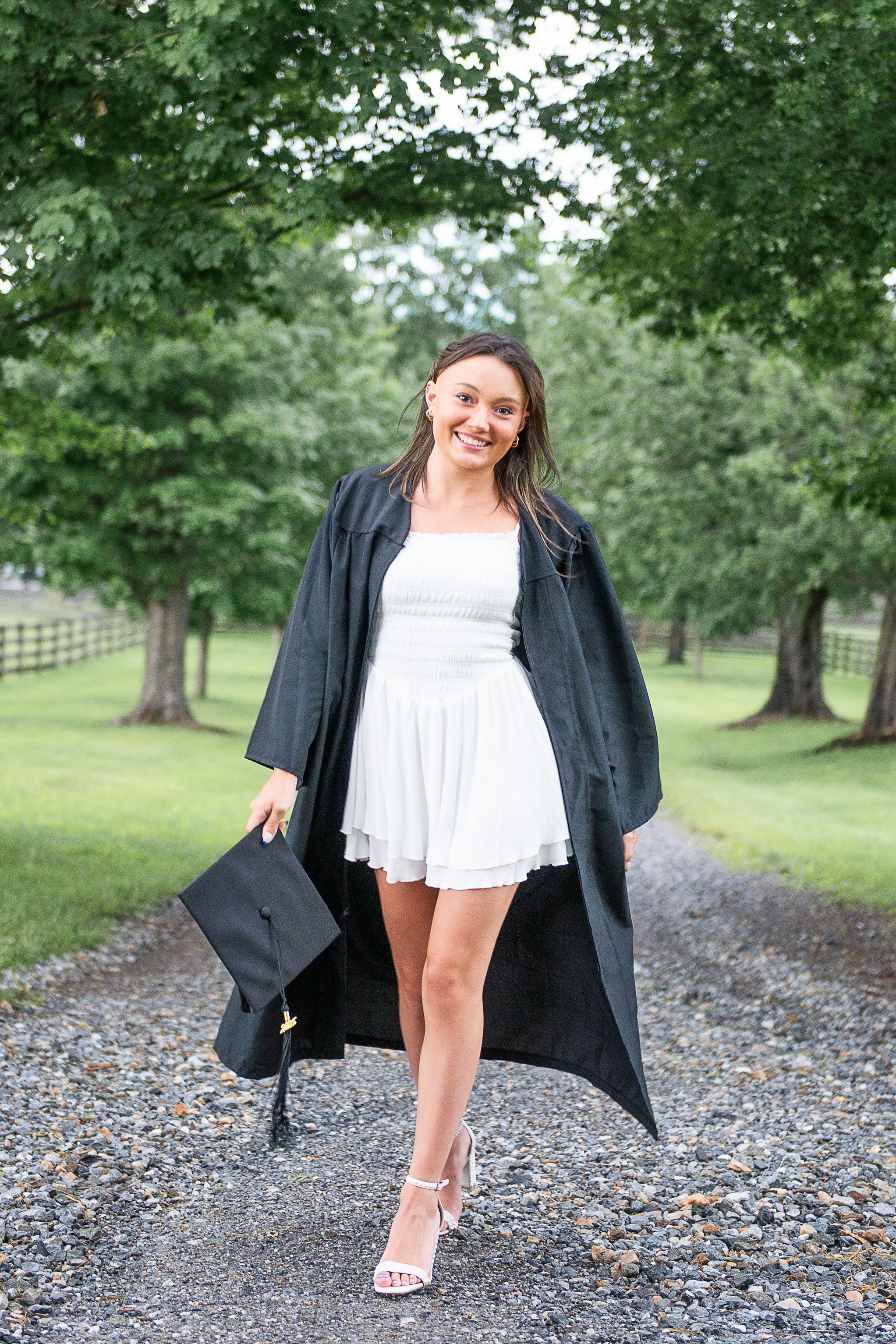 Graduation portrait showing a graduate in a white dress with black cap and gown walking on a tree lined path.