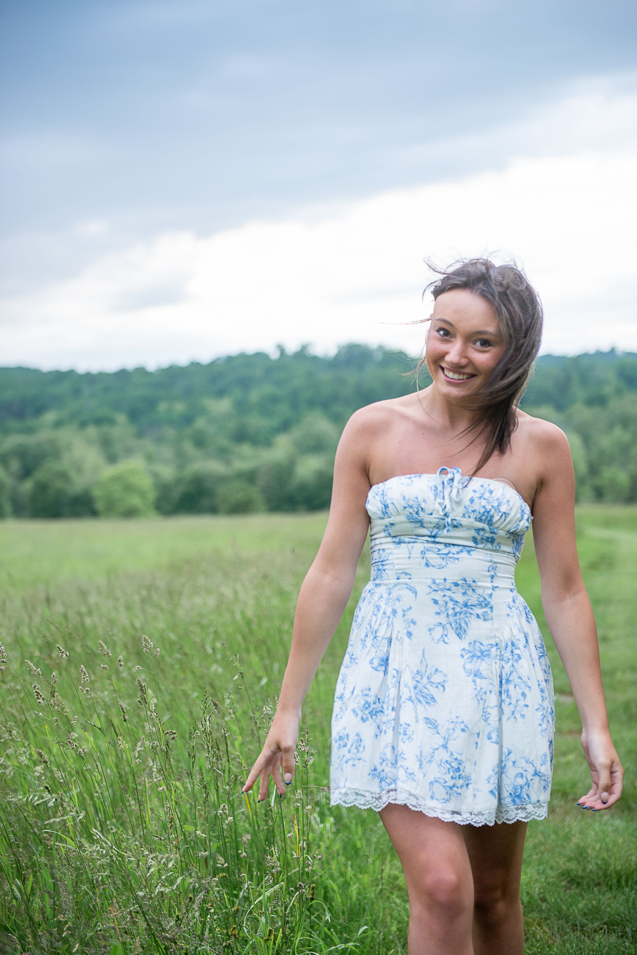 Playful poses in a blue and white floral sundress against lush green meadow background for a senior portrait.