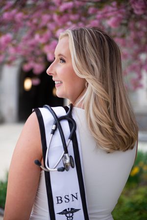 Nursing graduate in white dress with BSN stole smiling against pink blossoms.