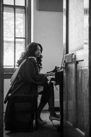 Black and white photo of a woman with curly hair sitting at an upright piano, glancing back toward the camera.
