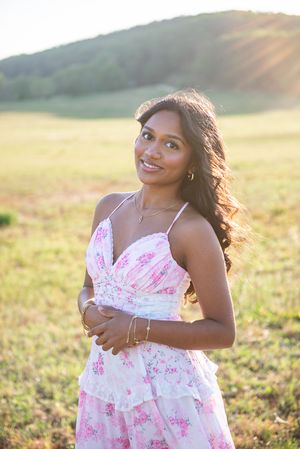 Young woman in pink floral sundress smiling in sunlit field with green hills in background.