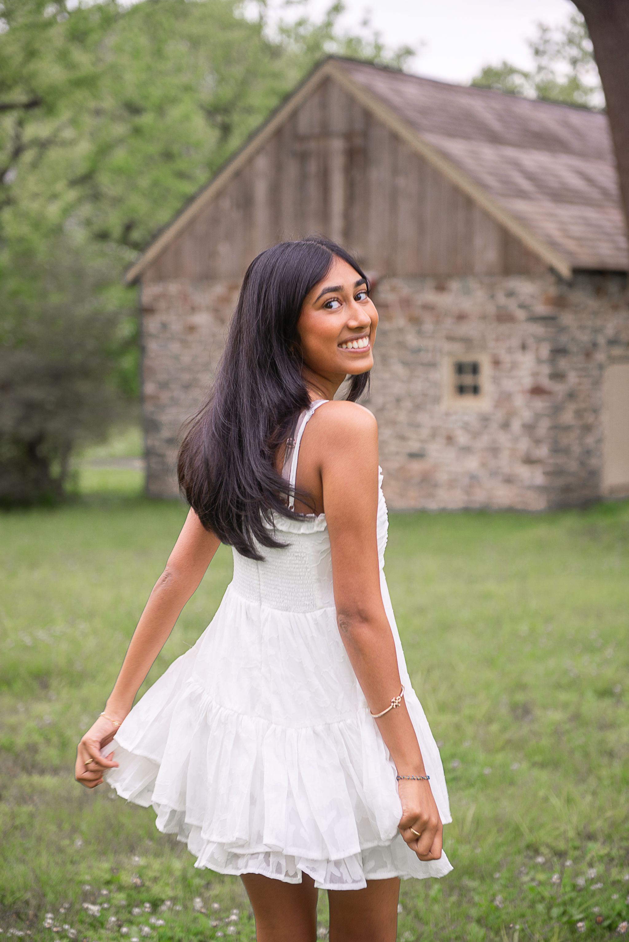 Young woman in white ruffled mini dress smiling over shoulder outdoors near rustic barn.