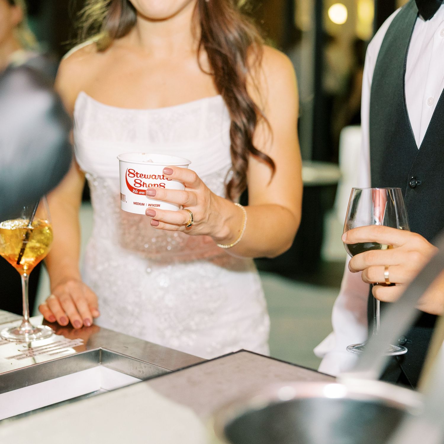 Person in white dress holding a drink at an event while bartender serves beverages at upscale bar.