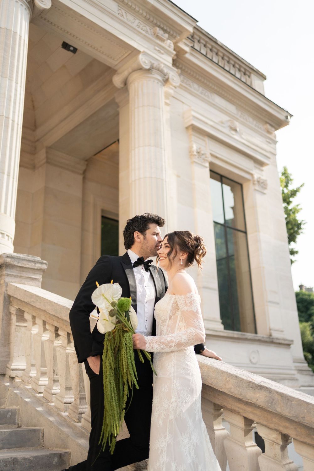 Elegant wedding couple poses on grand marble staircase with ornate columns at historic venue.