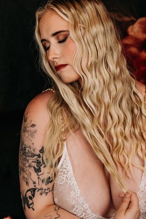 Artistic portrait shows blonde wavy hair, intricate arm tattoo, and white lace dress against dark background. Michigan Boudoir Photography