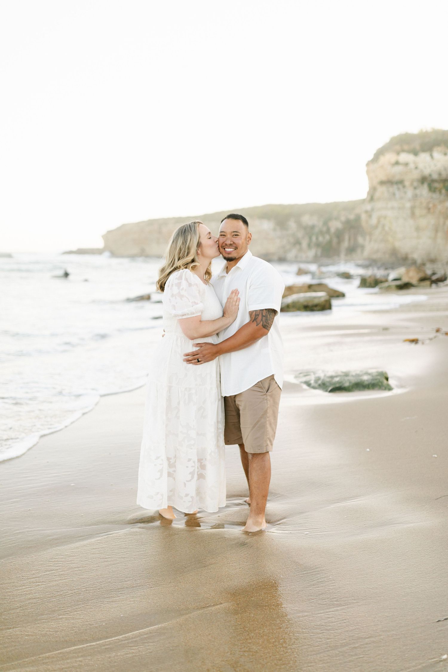 A romantic couple embraces on a sandy beach with dramatic coastal cliffs in the background during golden hour.