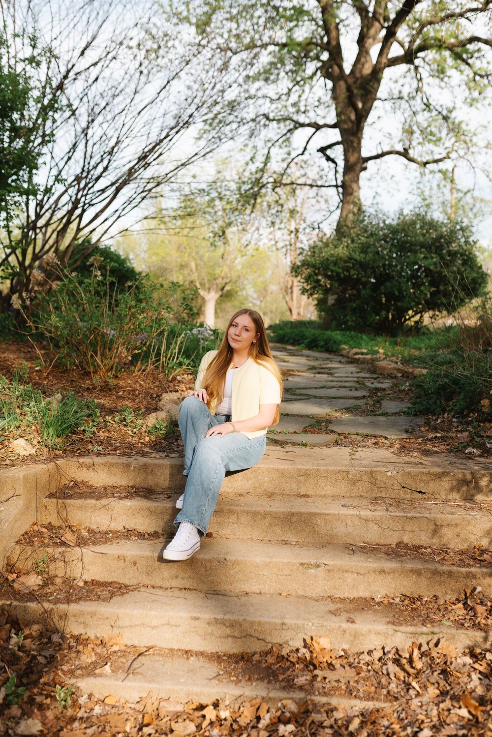 A person wearing light colored clothing sits on stone garden steps surrounded by trees and fallen leaves.