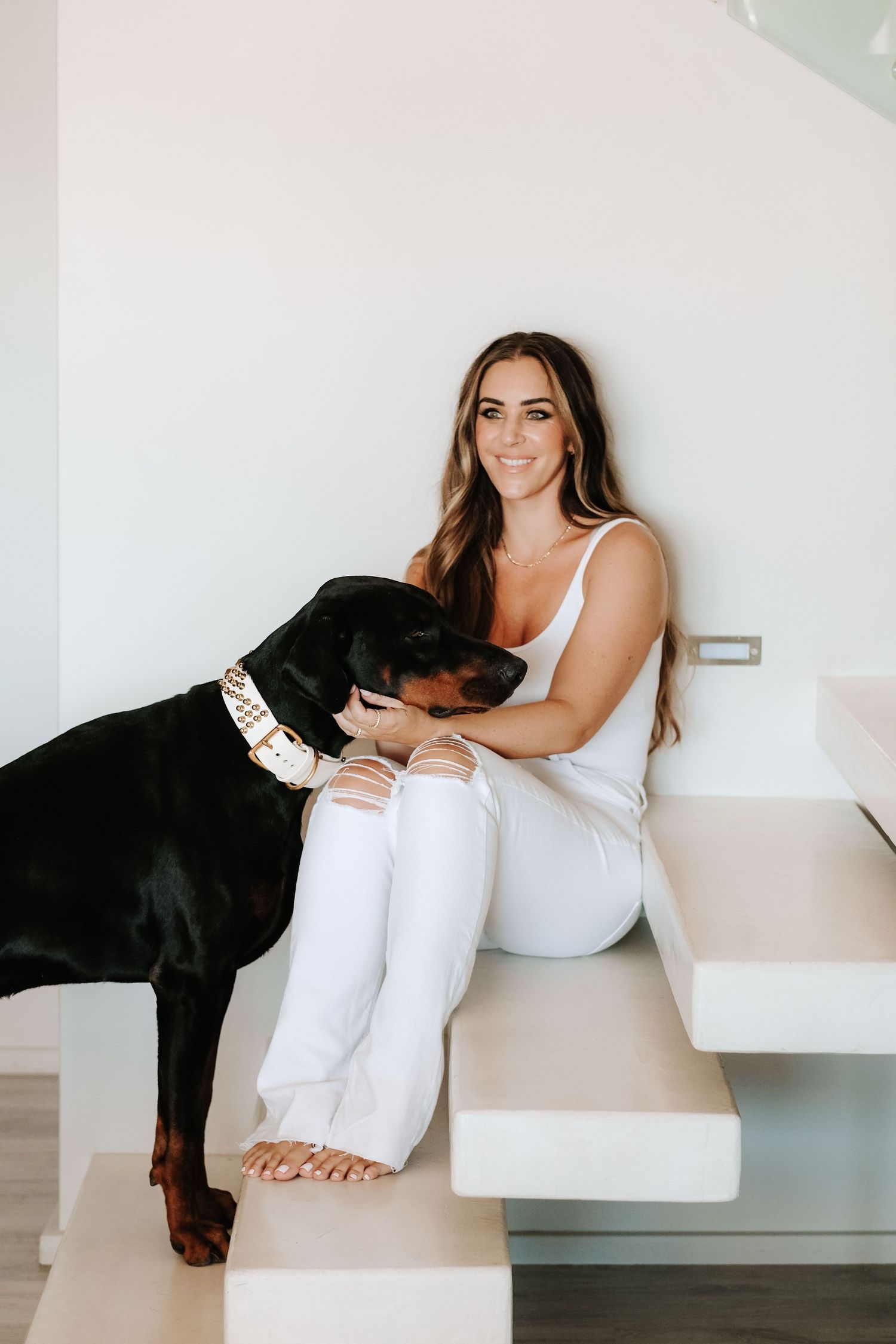 Smiling brunette woman in white outfit sits on stairs with black Doberman dog.