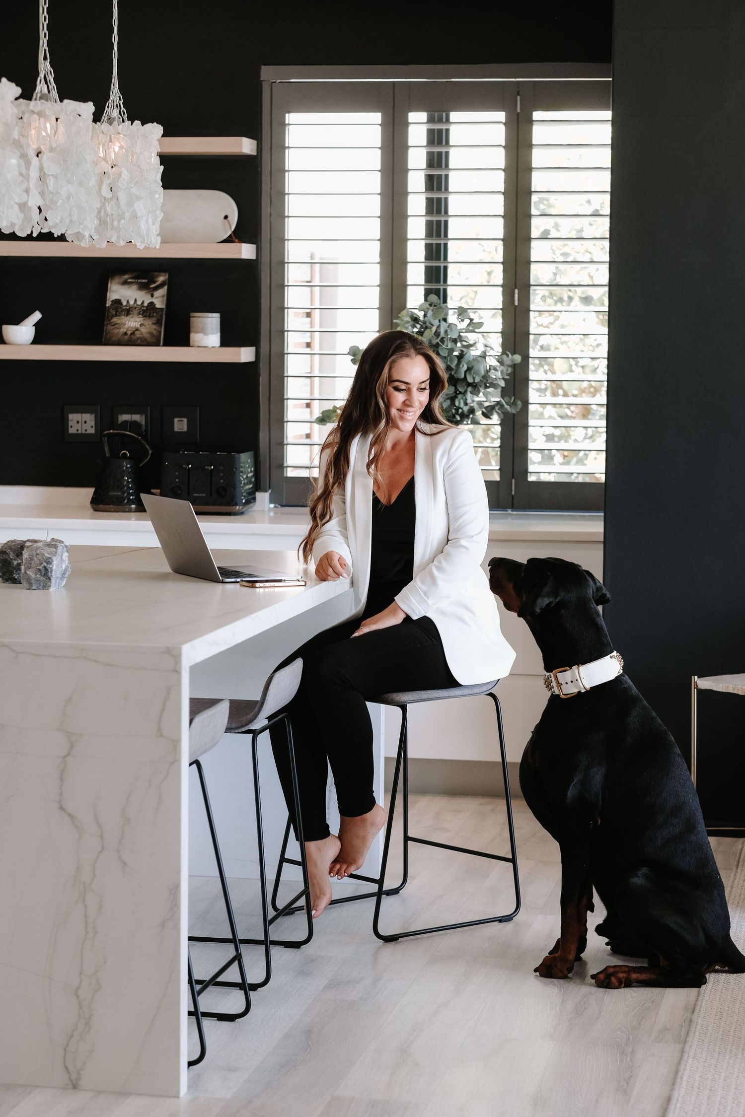 Smiling woman in white blazer works on laptop at marble kitchen island with dog.