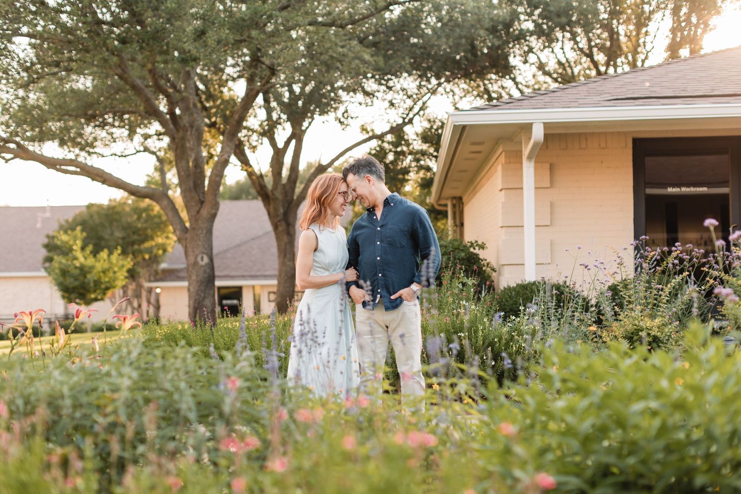 A couple shares an intimate moment in a garden at sunset with lush greenery and a cottage in the background.