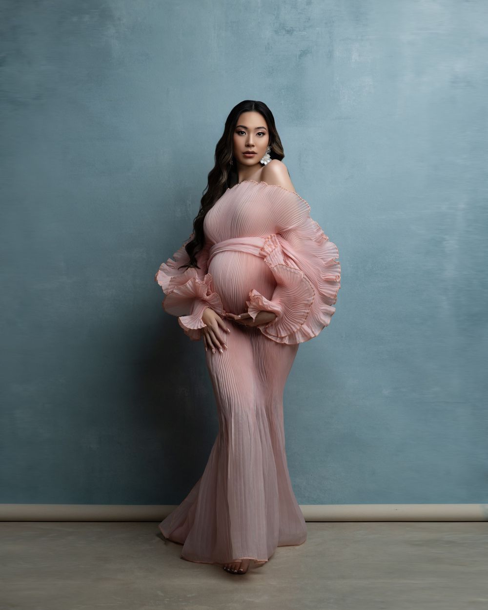 A pregnant figure poses in an elegant blush pink gown with dramatic ruffled details against a textured blue wall.