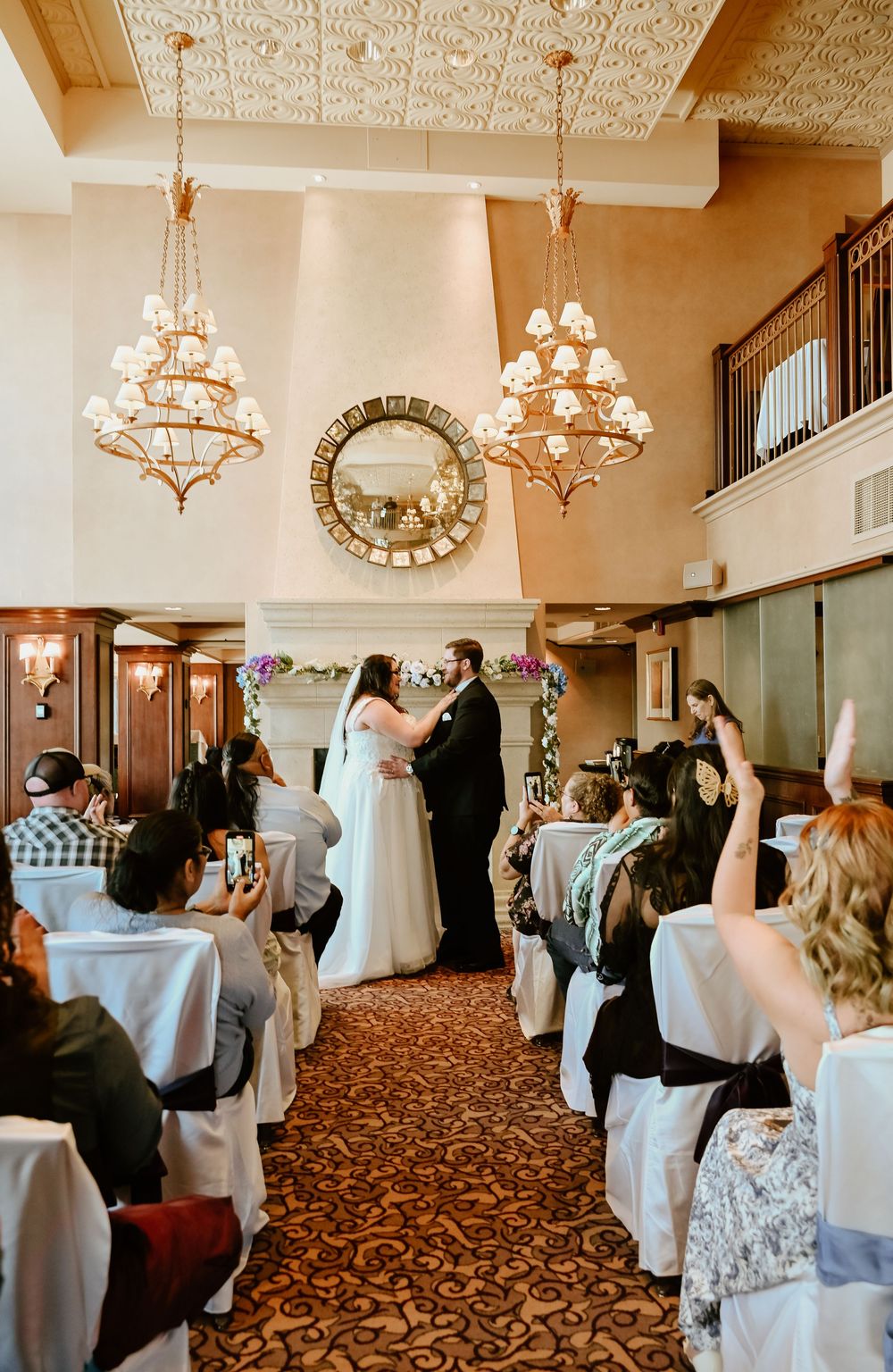 An elegant indoor wedding ceremony with ornate chandeliers, carpeted floors and white chair covers in a grand ballroom.