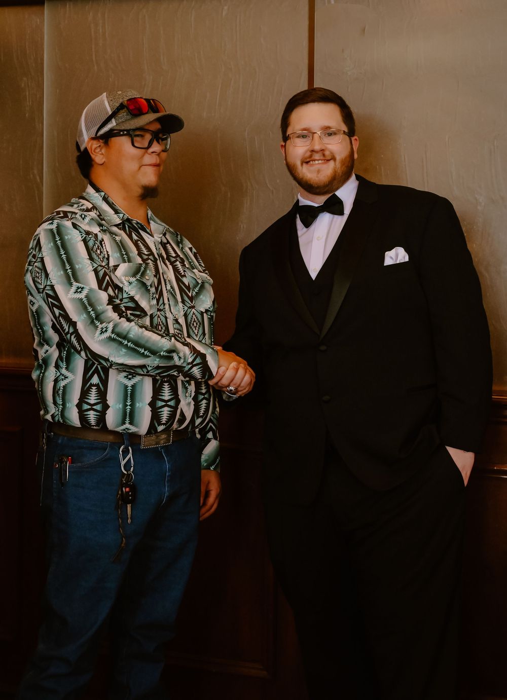 Two people shaking hands against a brown wall, one wearing casual attire and the other in a formal black tuxedo.