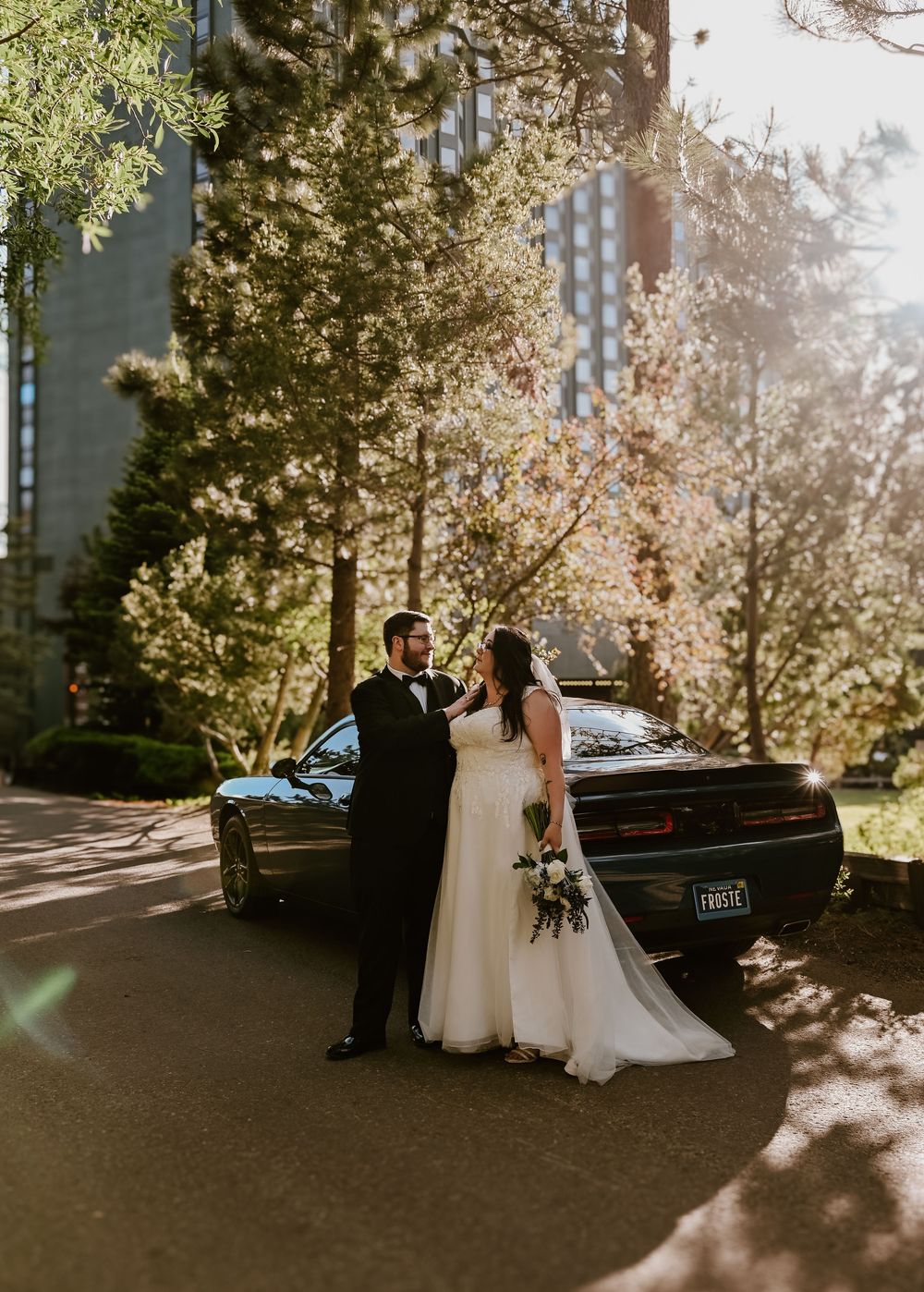 A wedding couple stands next to a black vintage car on a sunlit tree-lined street with golden rays filtering through.