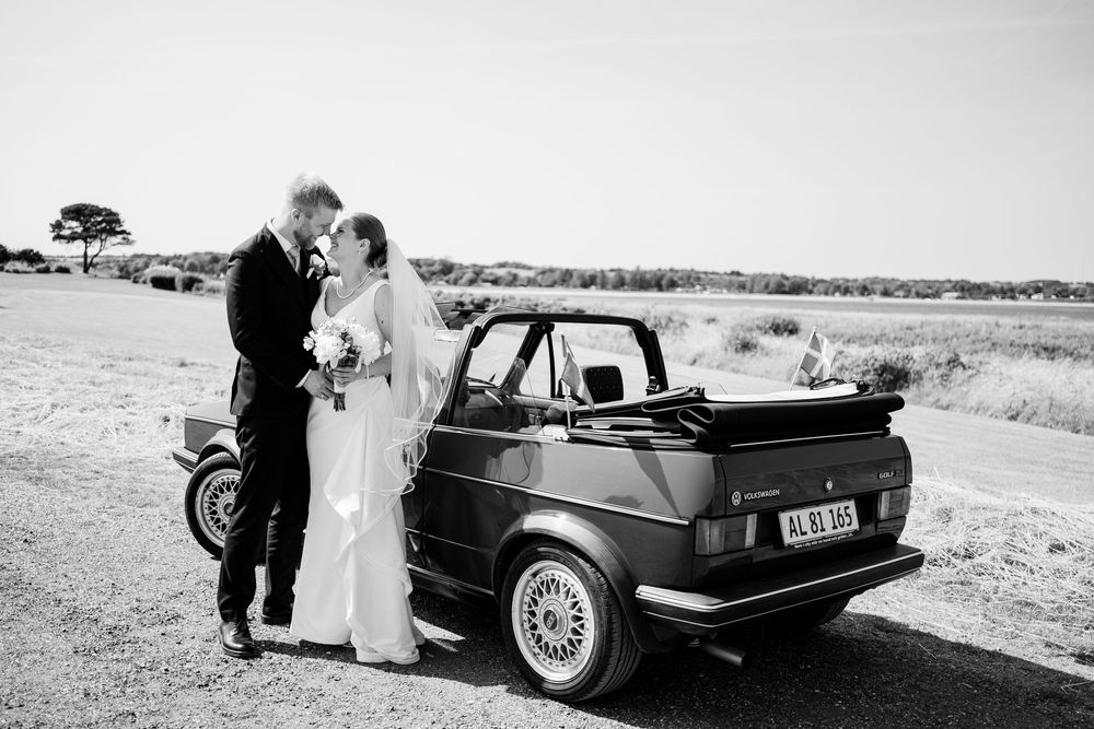 Classic vintage car parked in desert landscape with wedding party posing alongside in black and white photography.