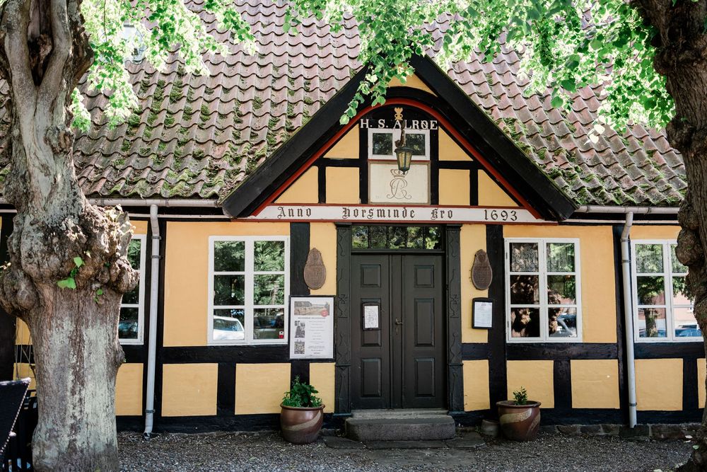 A charming yellow and black half-timbered Danish cottage with dark green door and tiled roof surrounded by trees.