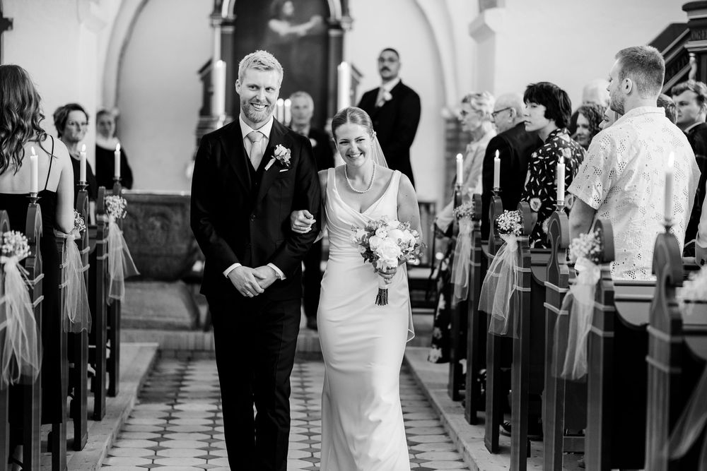 Black and white photo of newlyweds walking down the church aisle together as guests look on from the pews.