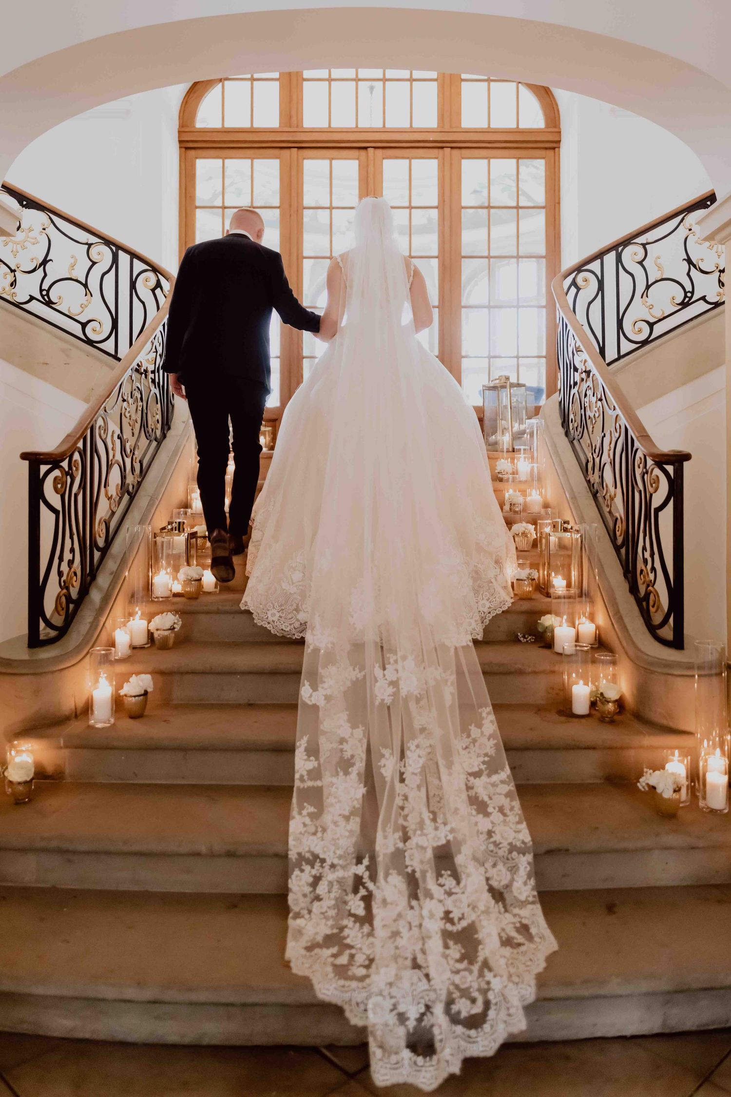 A couple ascends candlelit stairs with a flowing lace wedding dress train trailing behind them at a grand venue.
