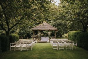Wooden gazebo set up for outdoor wedding ceremony with white chairs arranged on manicured lawn surrounded by trees.