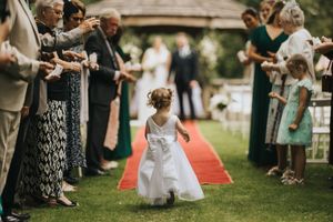 Young flower girl in white dress walks down red carpet aisle at outdoor garden wedding ceremony.