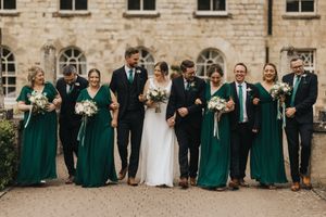 Wedding party in formal attire poses outdoors with emerald green bridesmaid dresses and black suits at stone building.