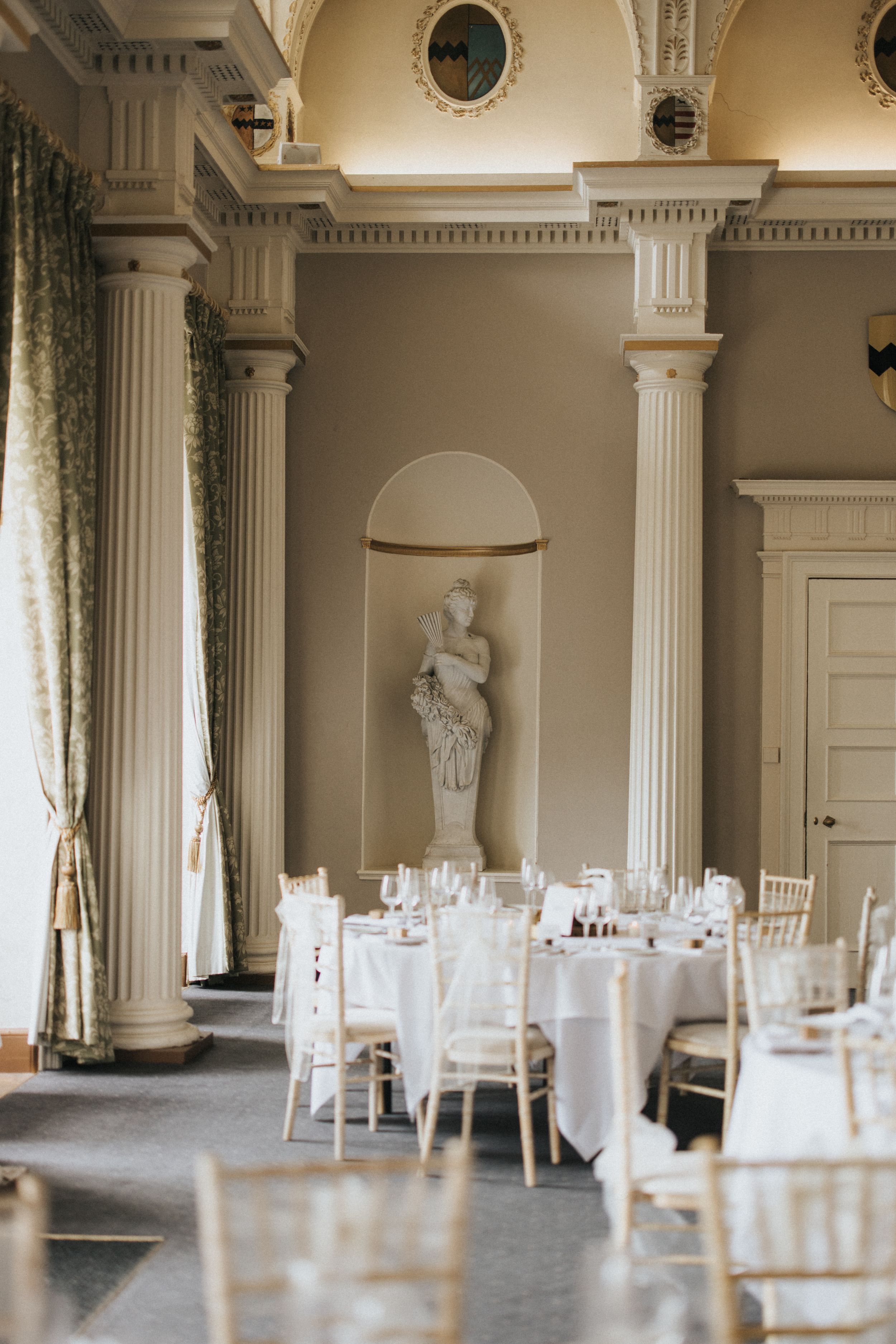 Elegant wedding venue with ornate white columns, draped windows, classical statue niche, and formal table settings.
