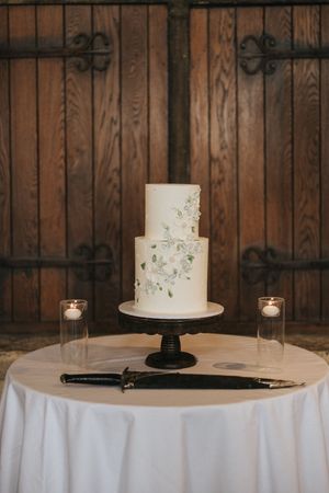 Elegant two-tier white wedding cake with delicate floral details displayed on black stand with candles on round table.