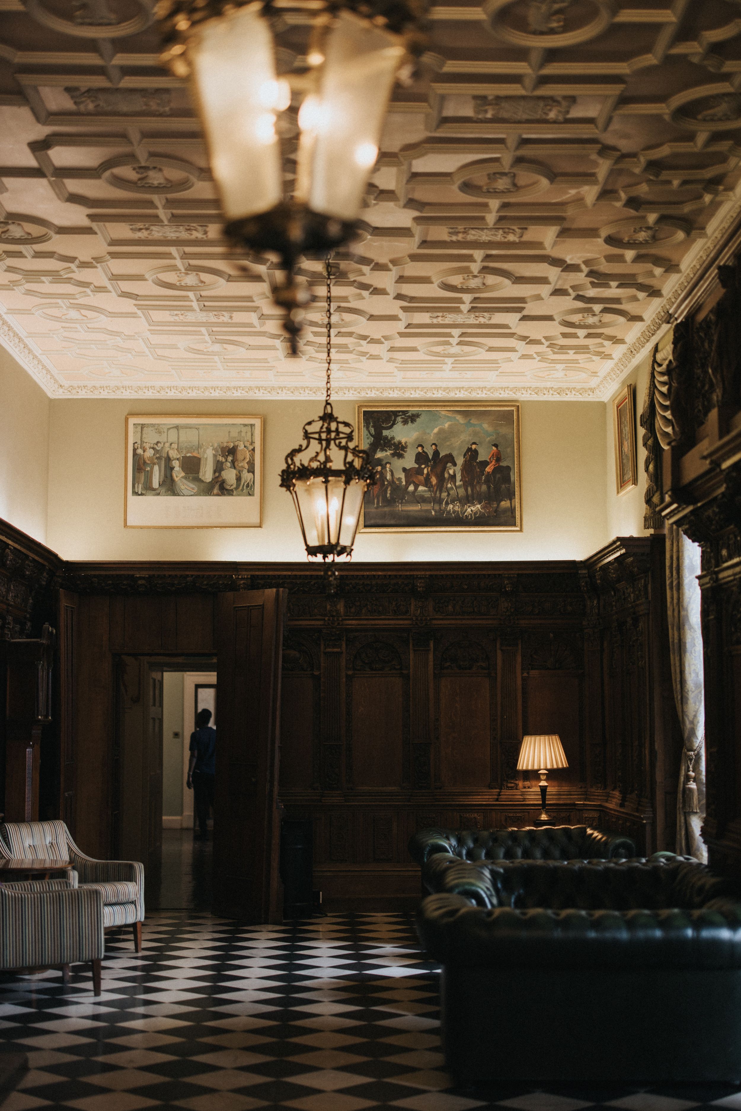 Dark vintage ballroom with ornate ceiling, black leather seating, and checkered floor illuminated by elegant lighting.