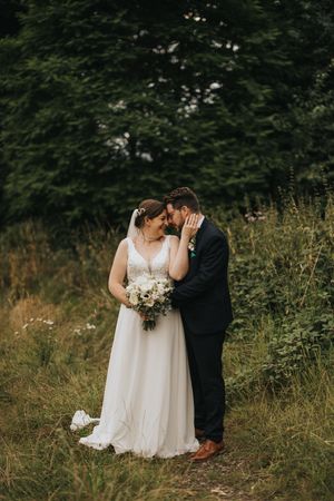 A romantic wedding couple embraces in a lush green meadow with natural outdoor lighting and wildflowers.