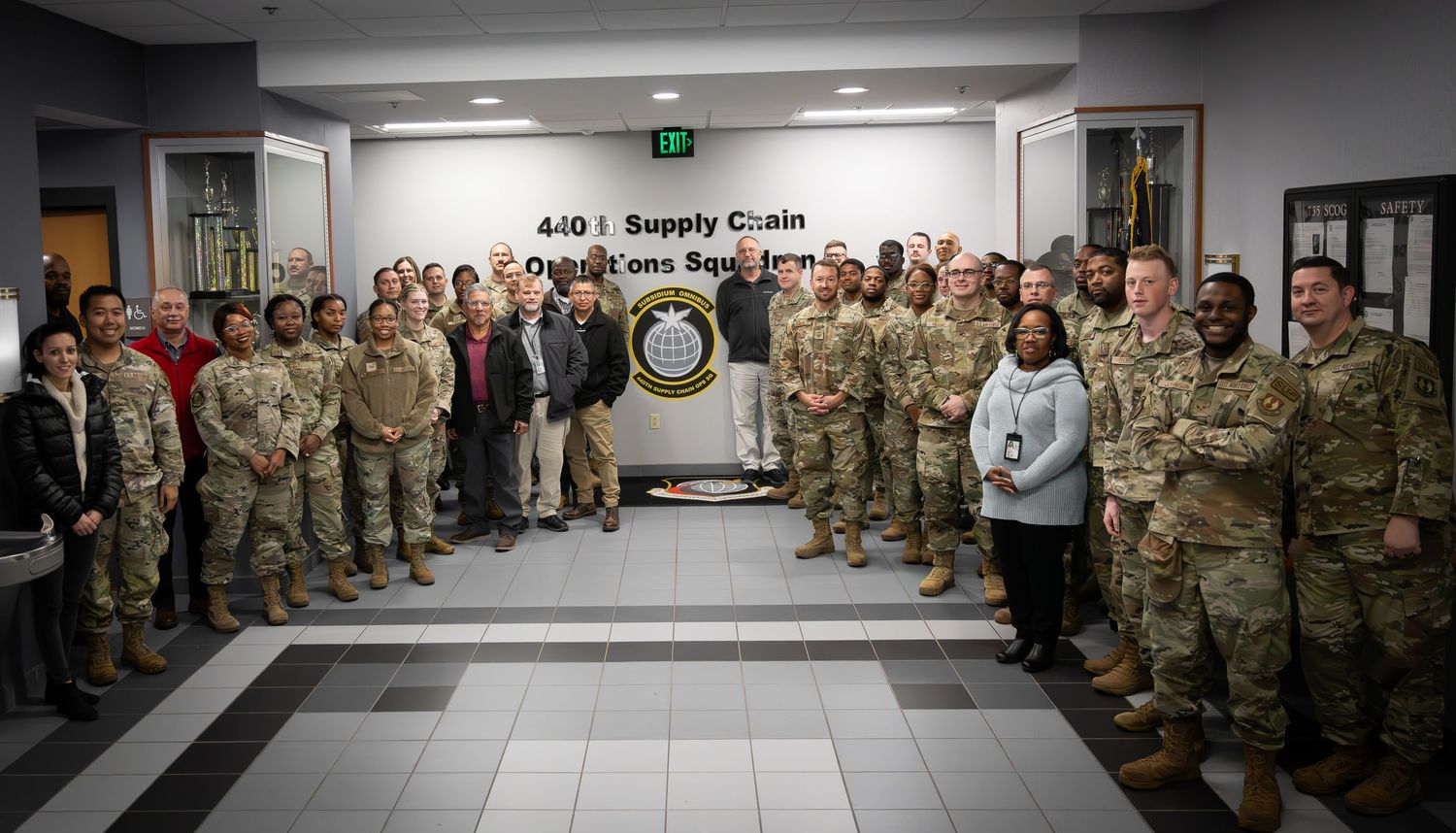 A large group of military personnel in camouflage uniforms stand together in a hallway at a supply circle facility.
