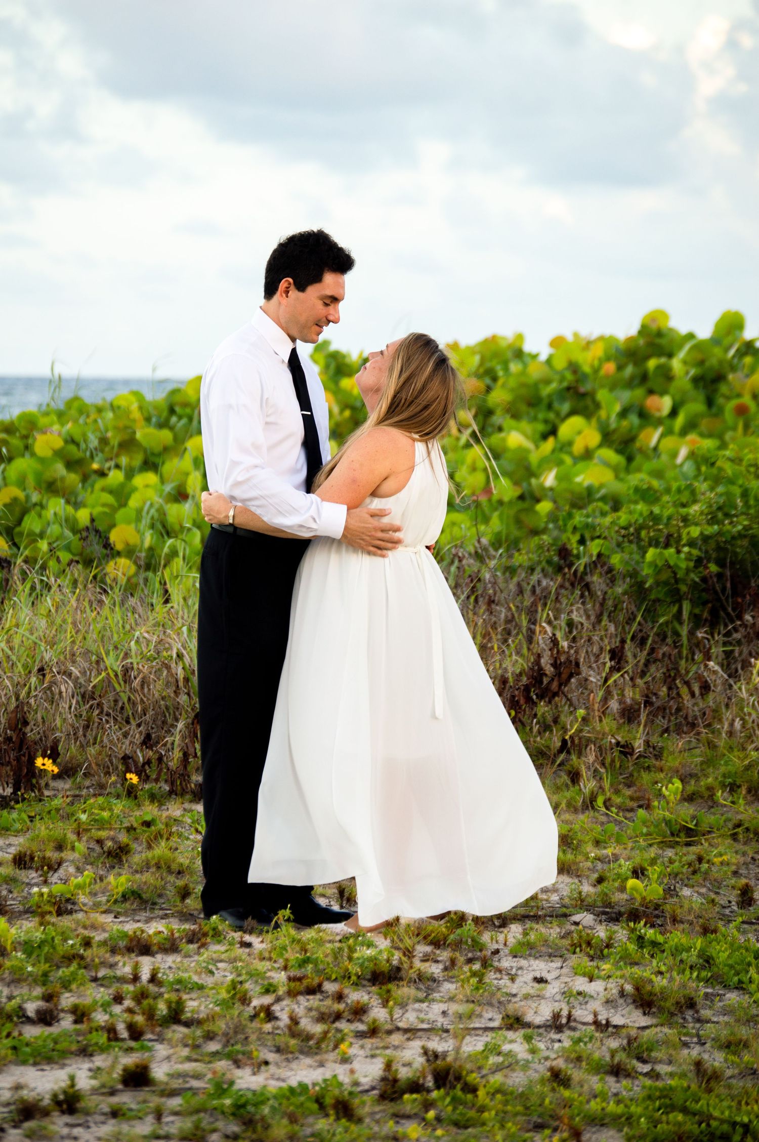 A couple in formal wedding attire has a romantic dance together in a lush green field by the coast.