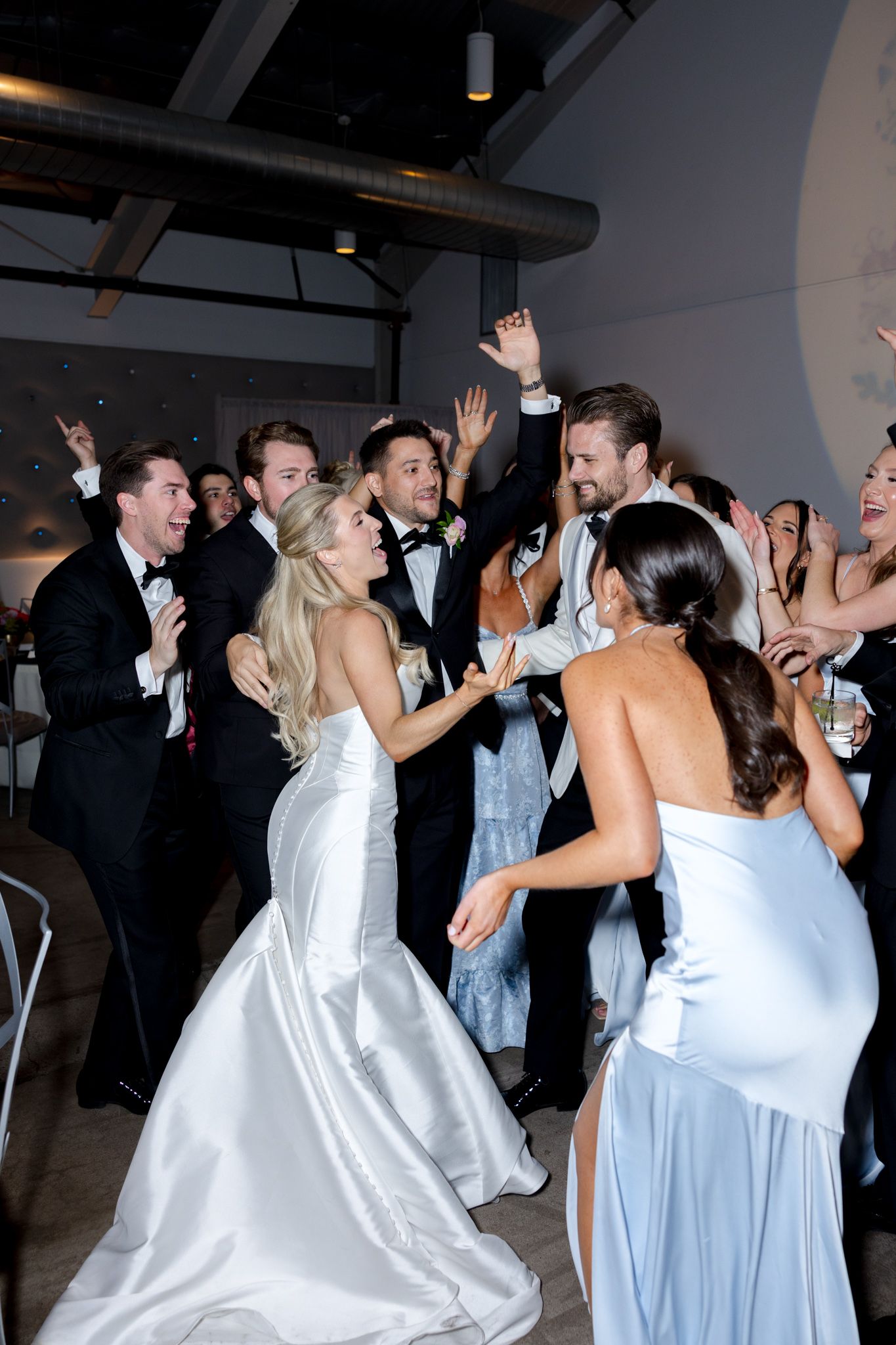 Wedding party celebrates on dance floor with bridesmaids in light blue dresses and men in formal attire.