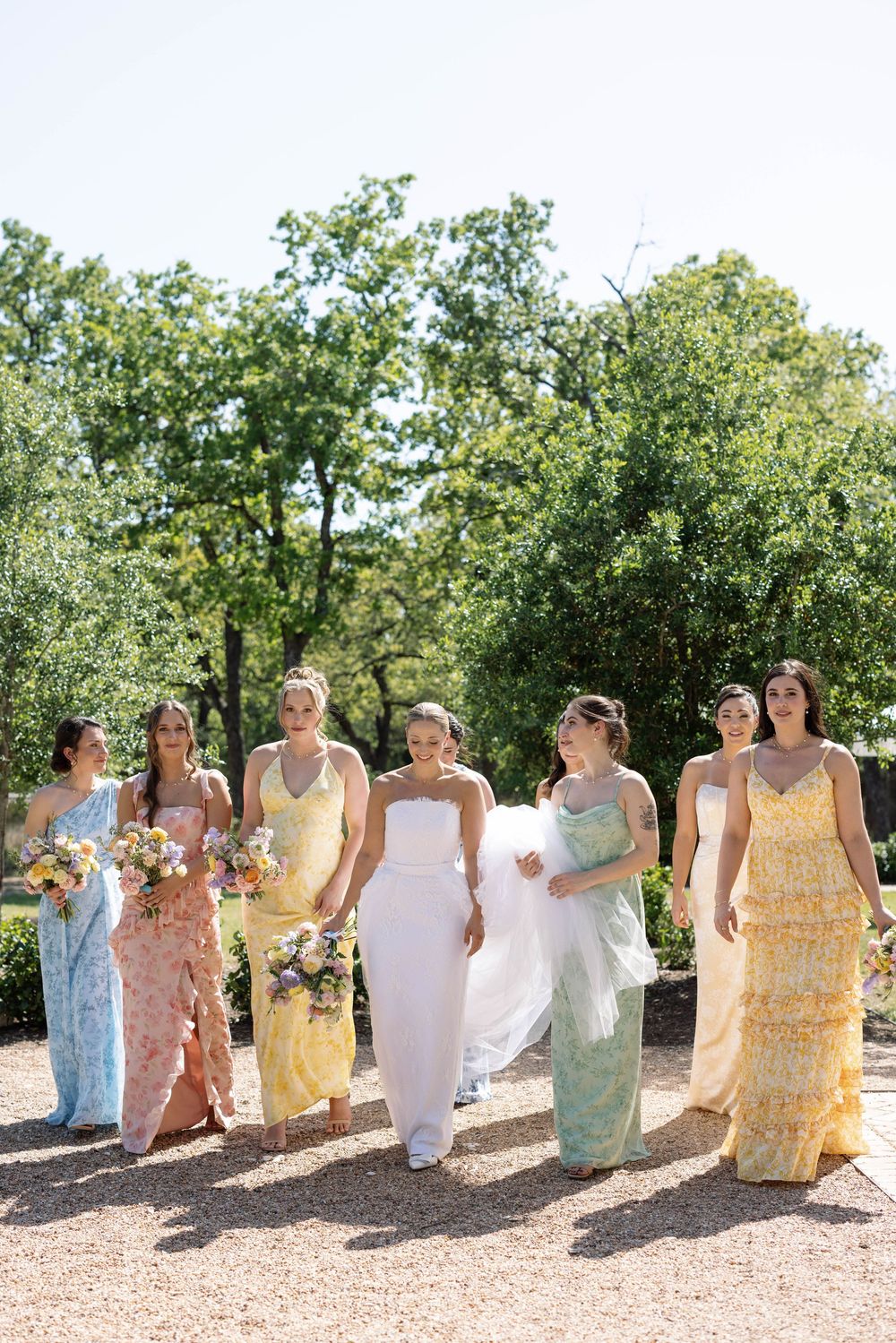 Bridal party wearing pastel floral dresses walks together outdoors on sunny day surrounded by green trees.