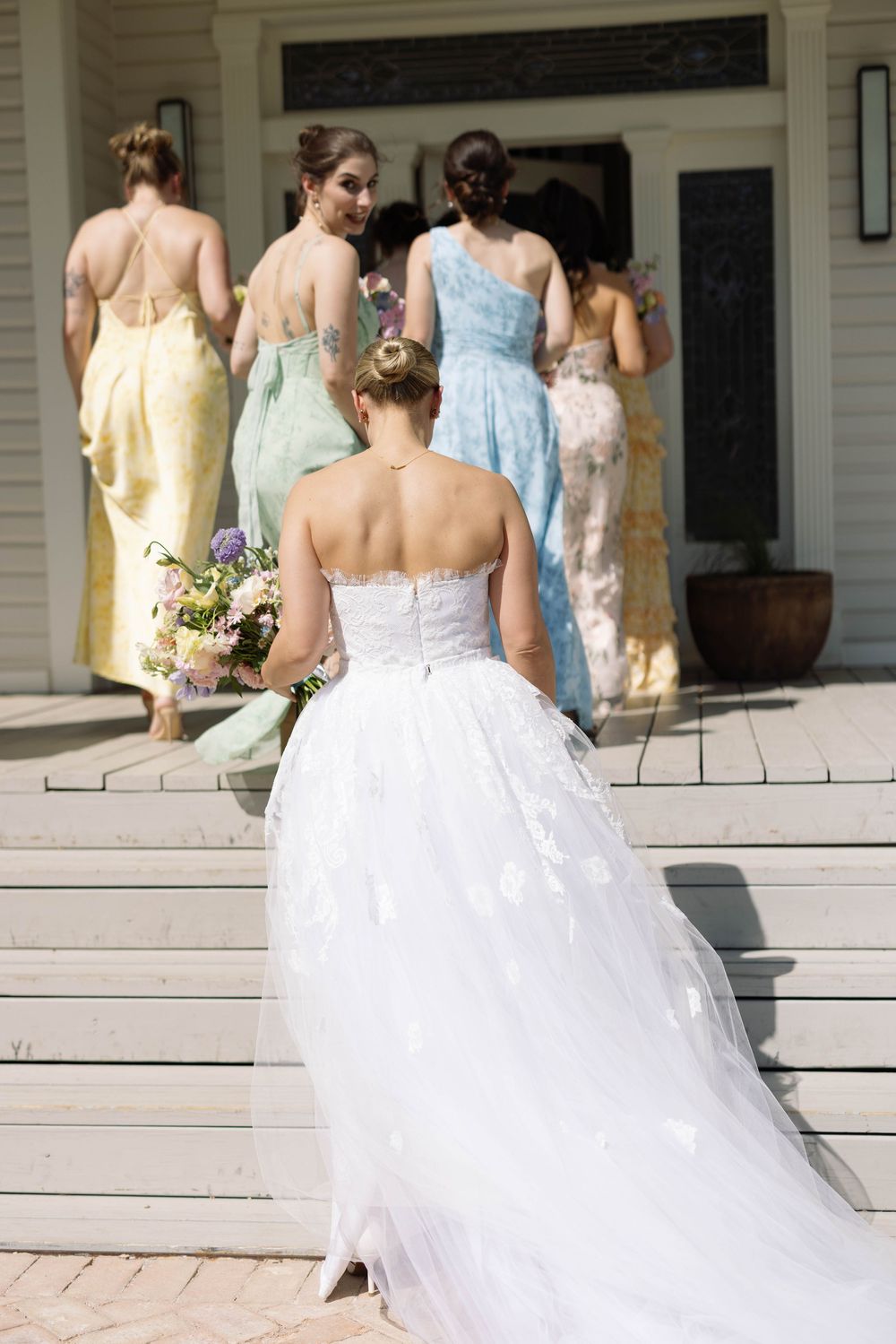 A group of bridesmaids in pastel dresses stand behind a bride in a white wedding gown on a sunny porch.