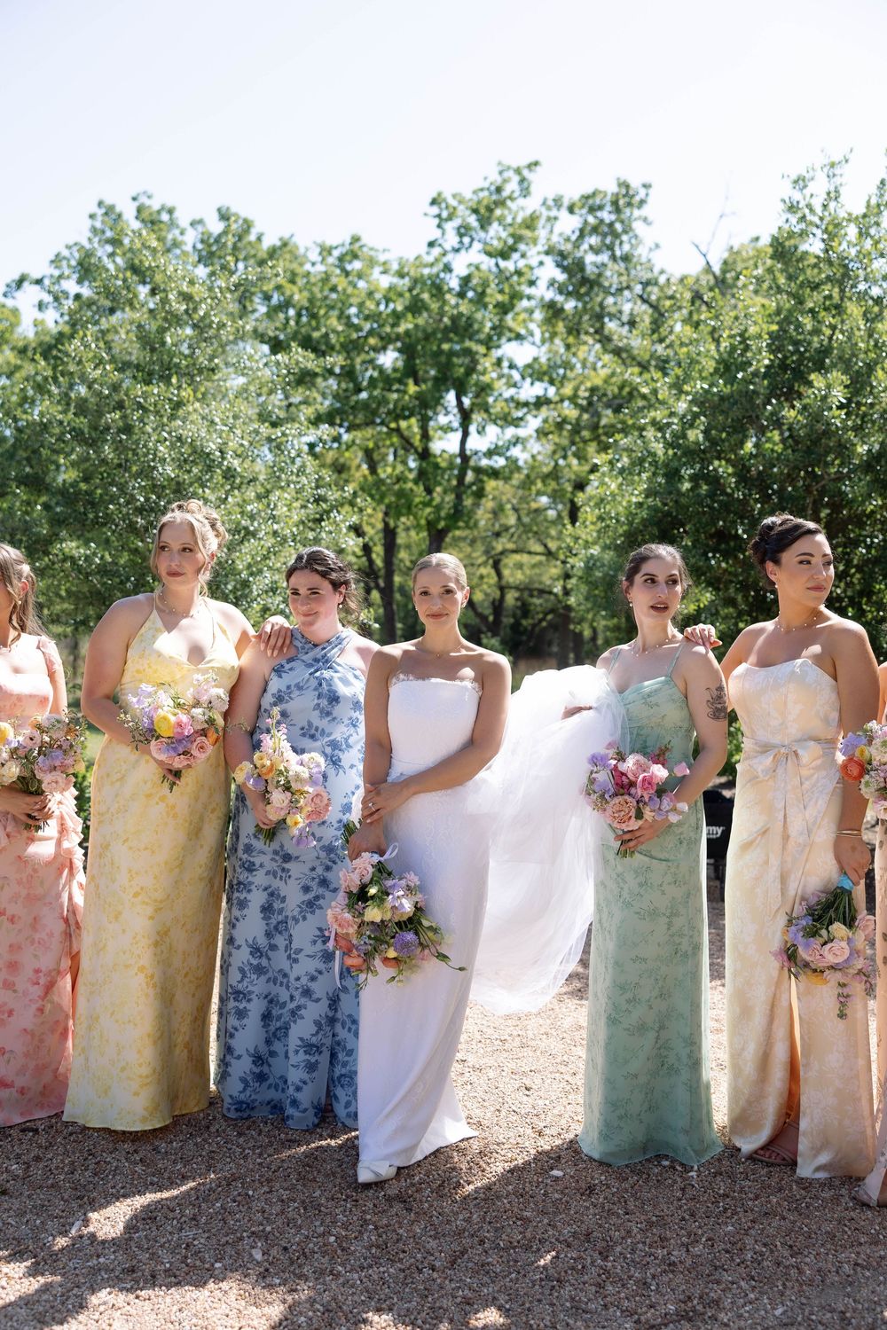 Bridal party stands outdoors in pastel floral dresses holding colorful bouquets on a sunny summer day.