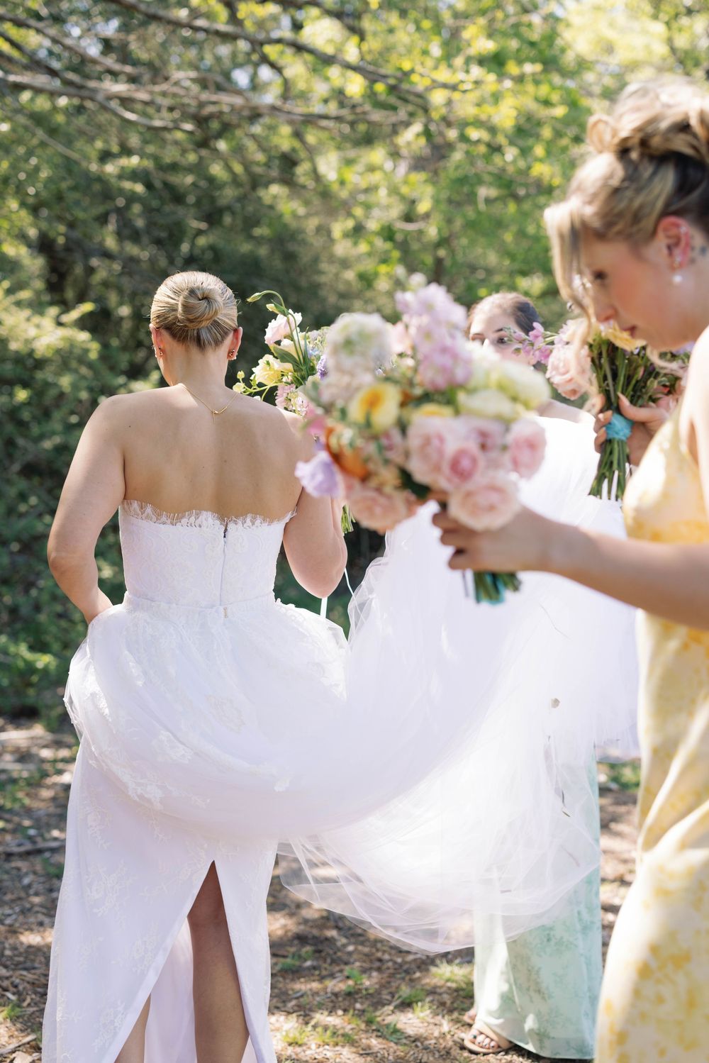 Bridesmaids in pastel yellow dresses hold bouquets while walking outdoors alongside bride in white wedding gown.