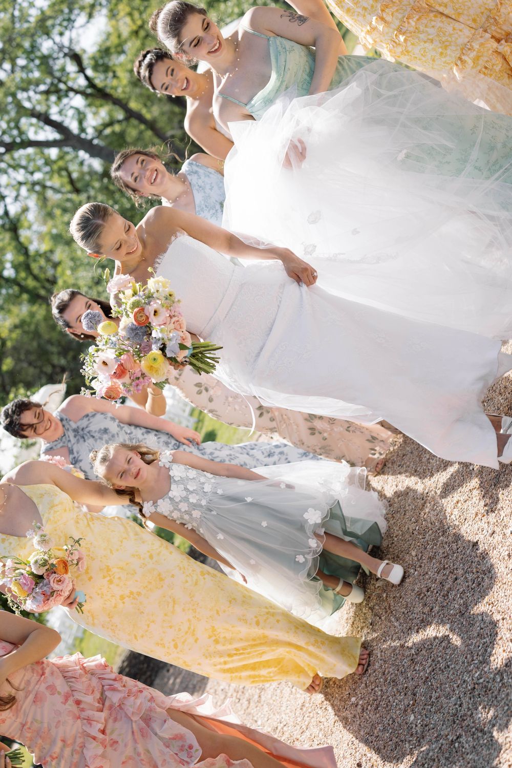 A wedding party in pastel dresses stands outside on a sunny day holding colorful floral bouquets.