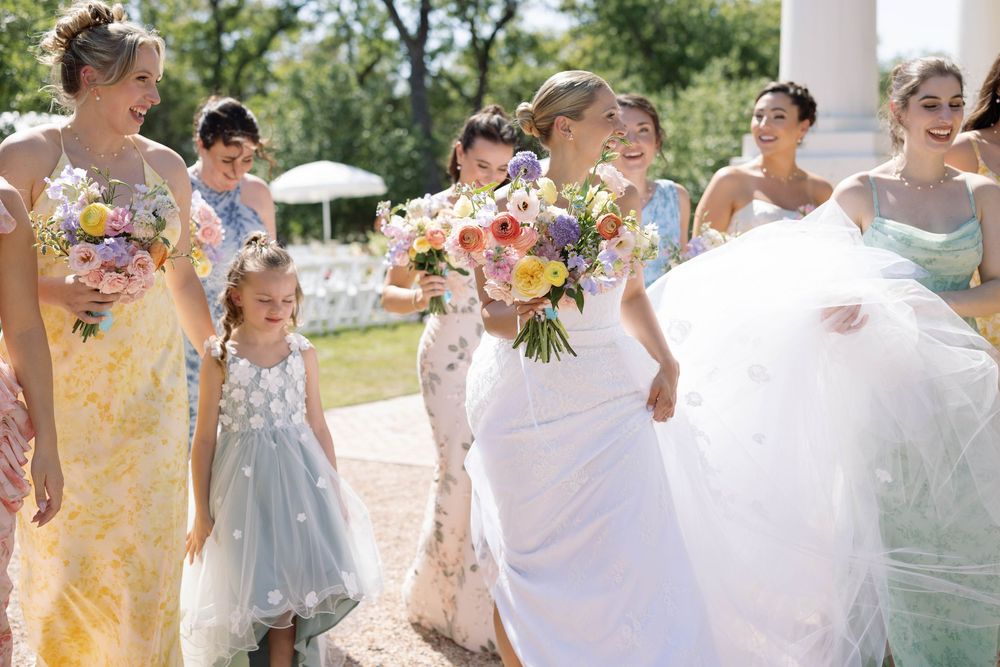 A bridal party in light colored dresses and suits walks together outside on a sunny summer day.