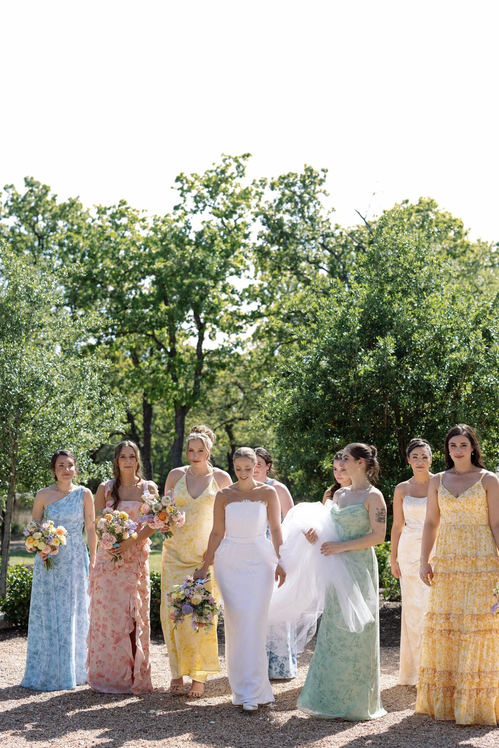 A group wearing summer dresses in pastel colors stands posed together outdoors on a sunny day.