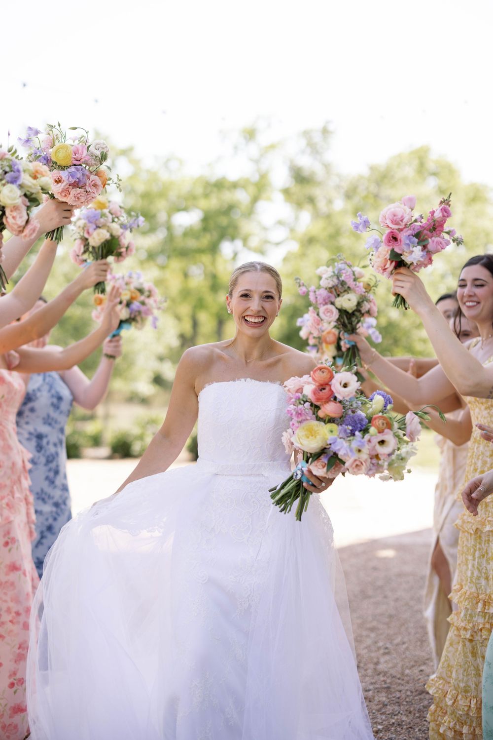 Beautiful wedding party holds up colorful floral bouquets while celebrating outdoors on a sunny day.