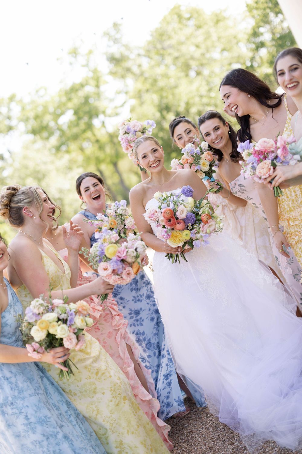 A bridal party wearing pastel floral dresses laughs together while holding colorful spring bouquets outdoors.