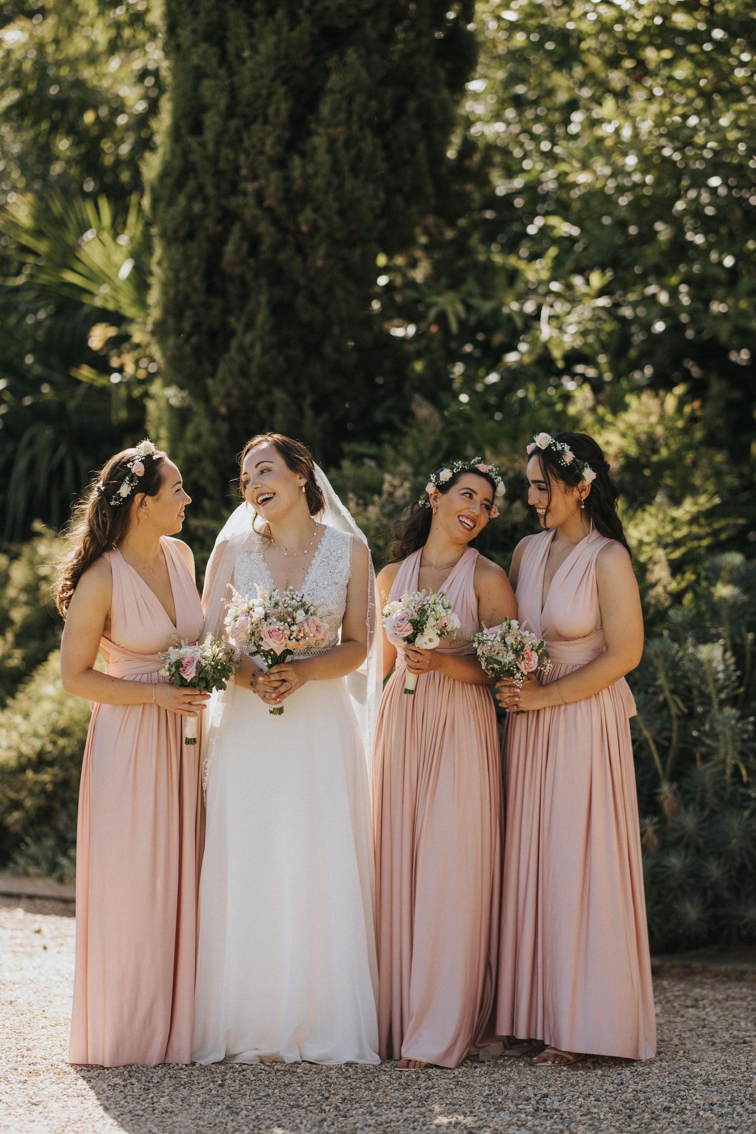 A bride in white gown stands with bridesmaids wearing blush pink dresses outdoors during a wedding photoshoot.