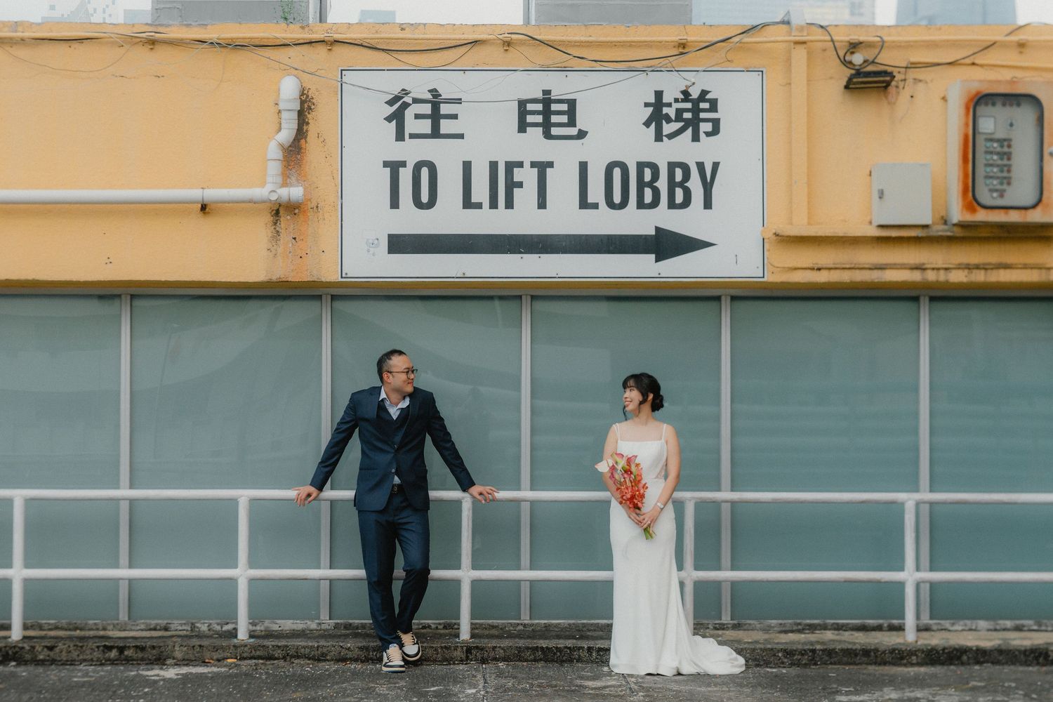A couple in wedding attire stands casually against a wall with Chinese and English directional signage to the lift lobby.