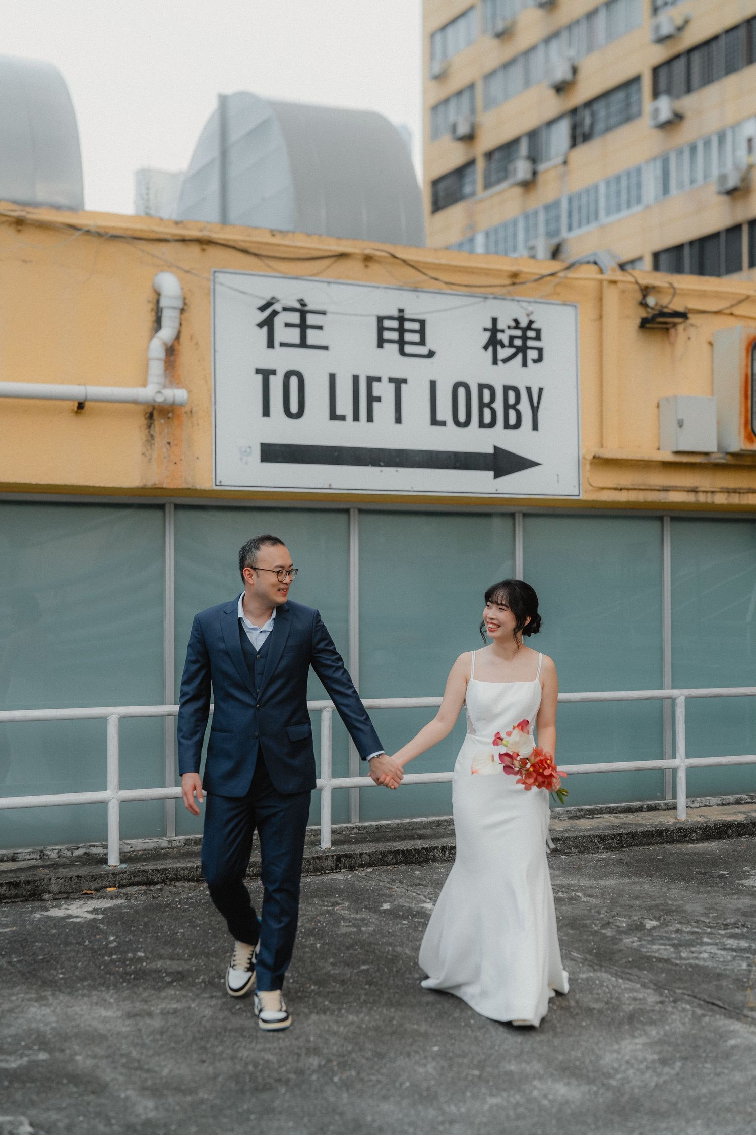 A couple in wedding attire poses playfully near a rooftop elevator lobby sign in an urban setting.