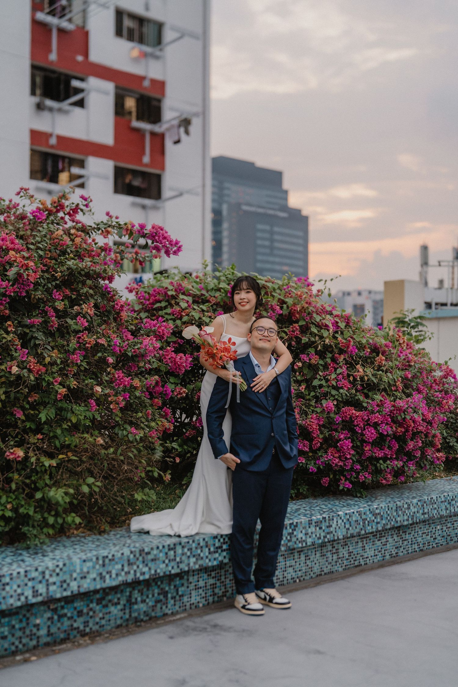 A couple posing against vibrant pink bougainvillea flowers with urban buildings in the background during sunset.