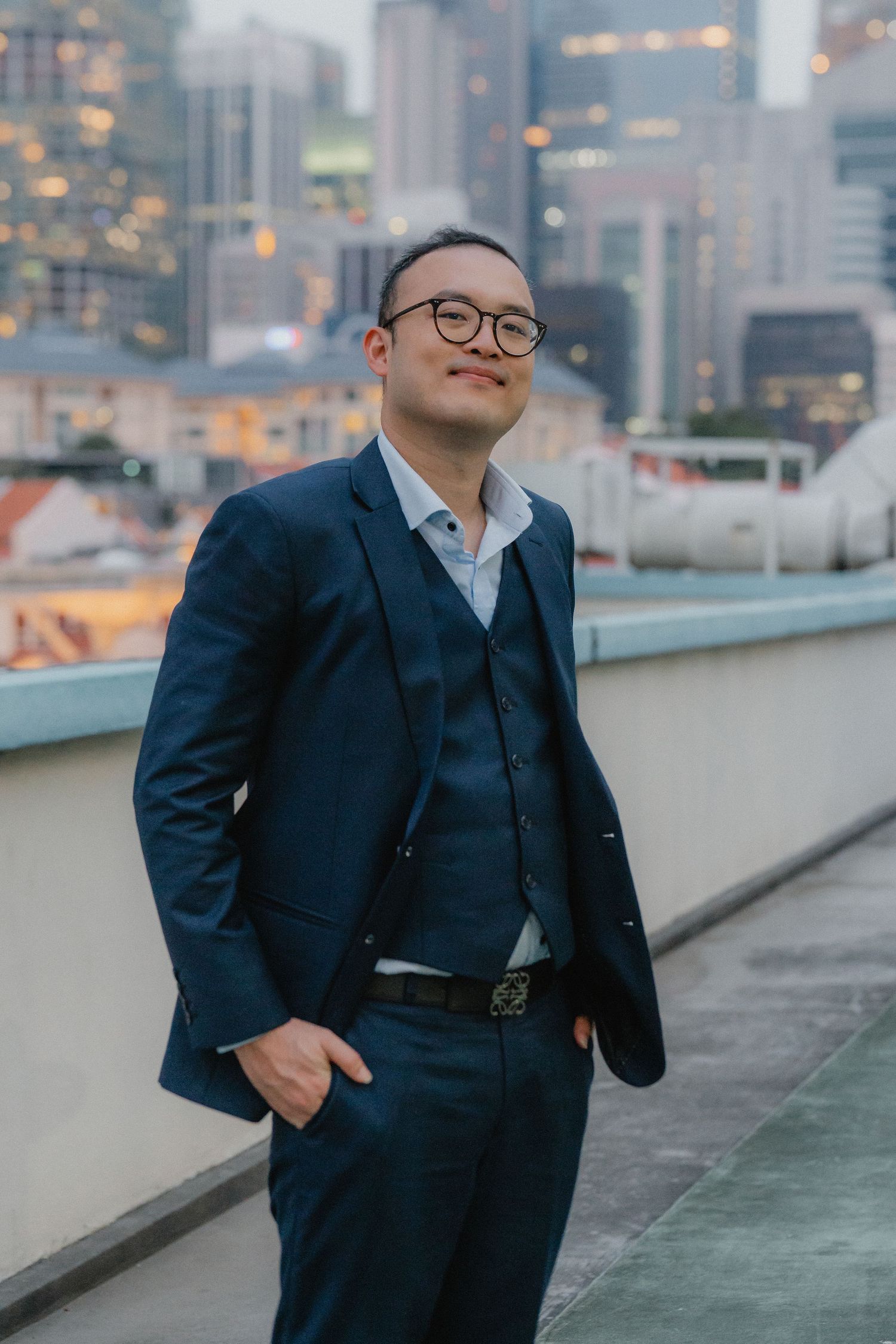 Professional looking man in a navy three-piece suit poses confidently on a rooftop with city buildings behind.