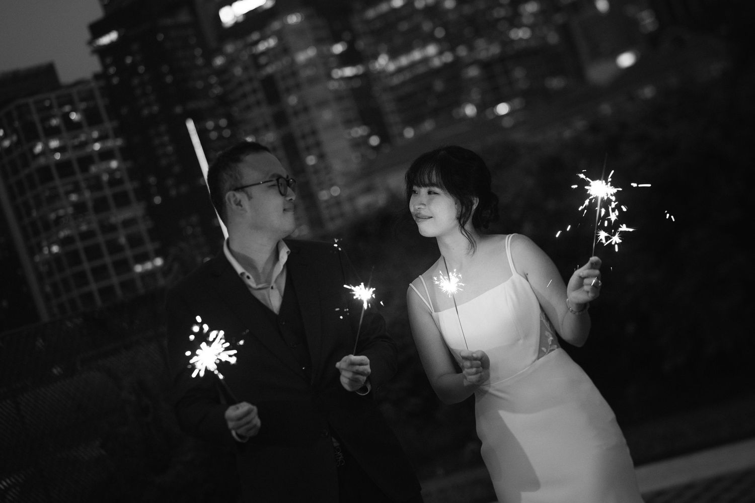 A couple in formal attire holds sparklers at night with a city skyline in the background.