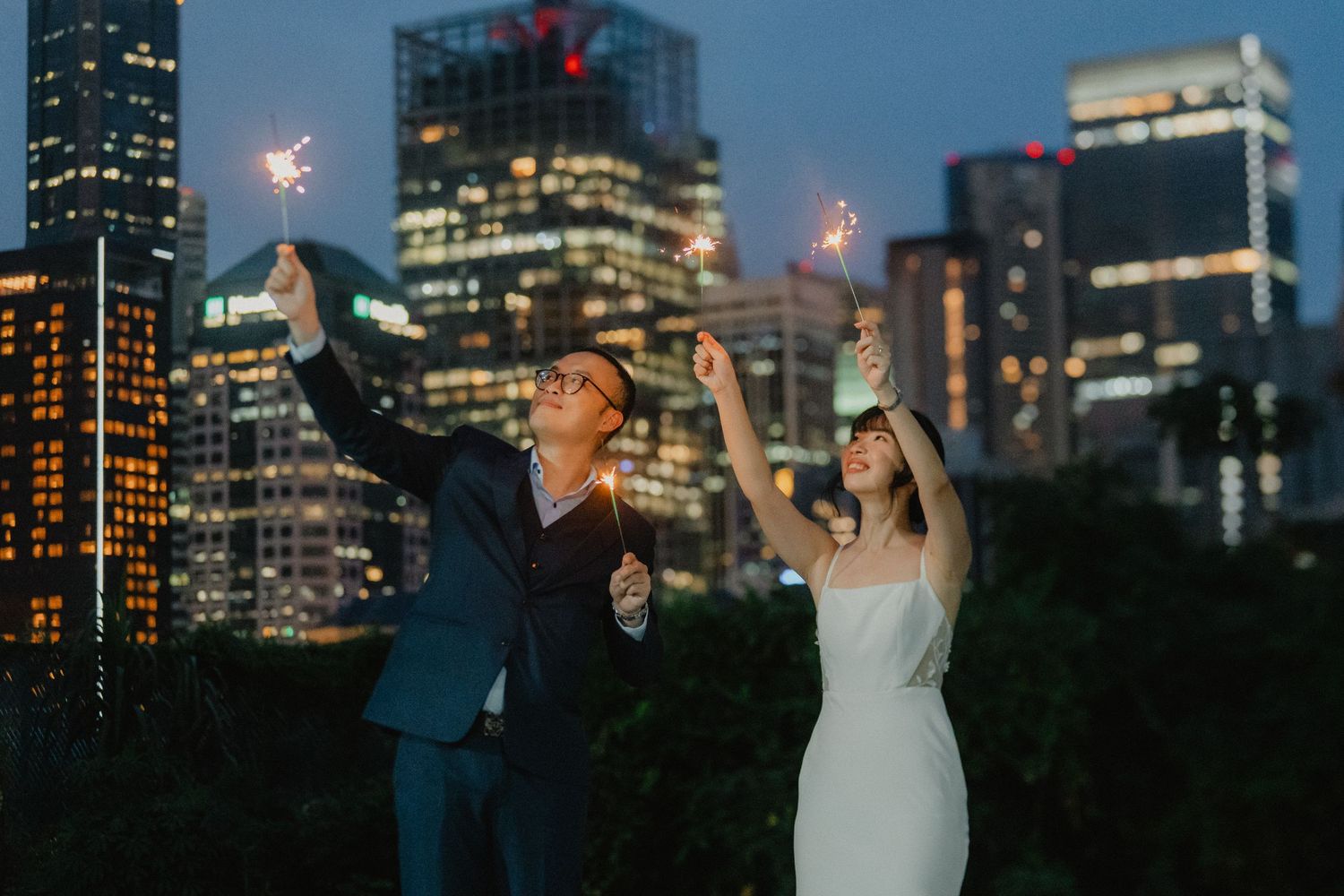 A couple celebrates with sparklers against a backdrop of illuminated city skyscrapers at twilight.