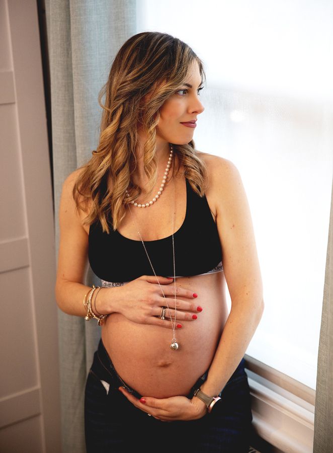 A pregnant woman in a black top stands by a window wearing pearl necklaces and bracelets while cradling her baby bump during a photoshoot