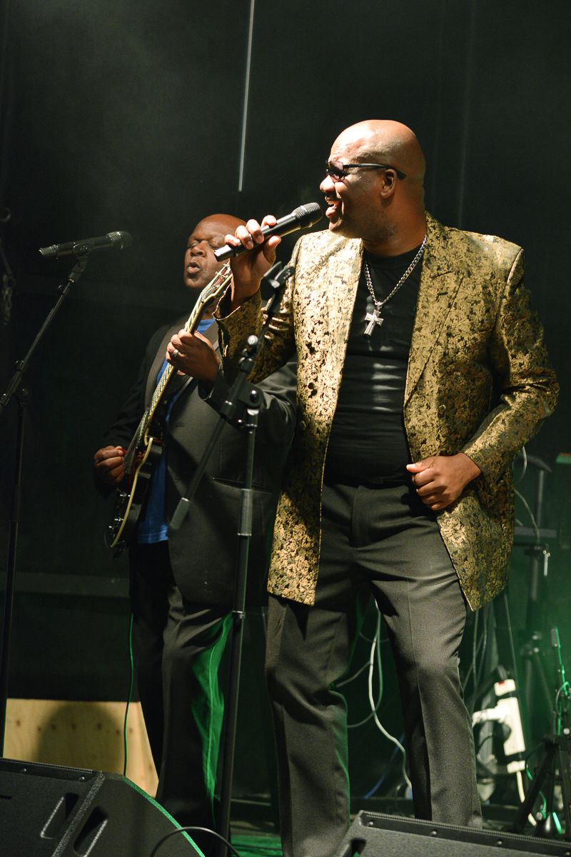 Performers on stage during a concert, with one playing guitar and another singing in a gold patterned blazer.